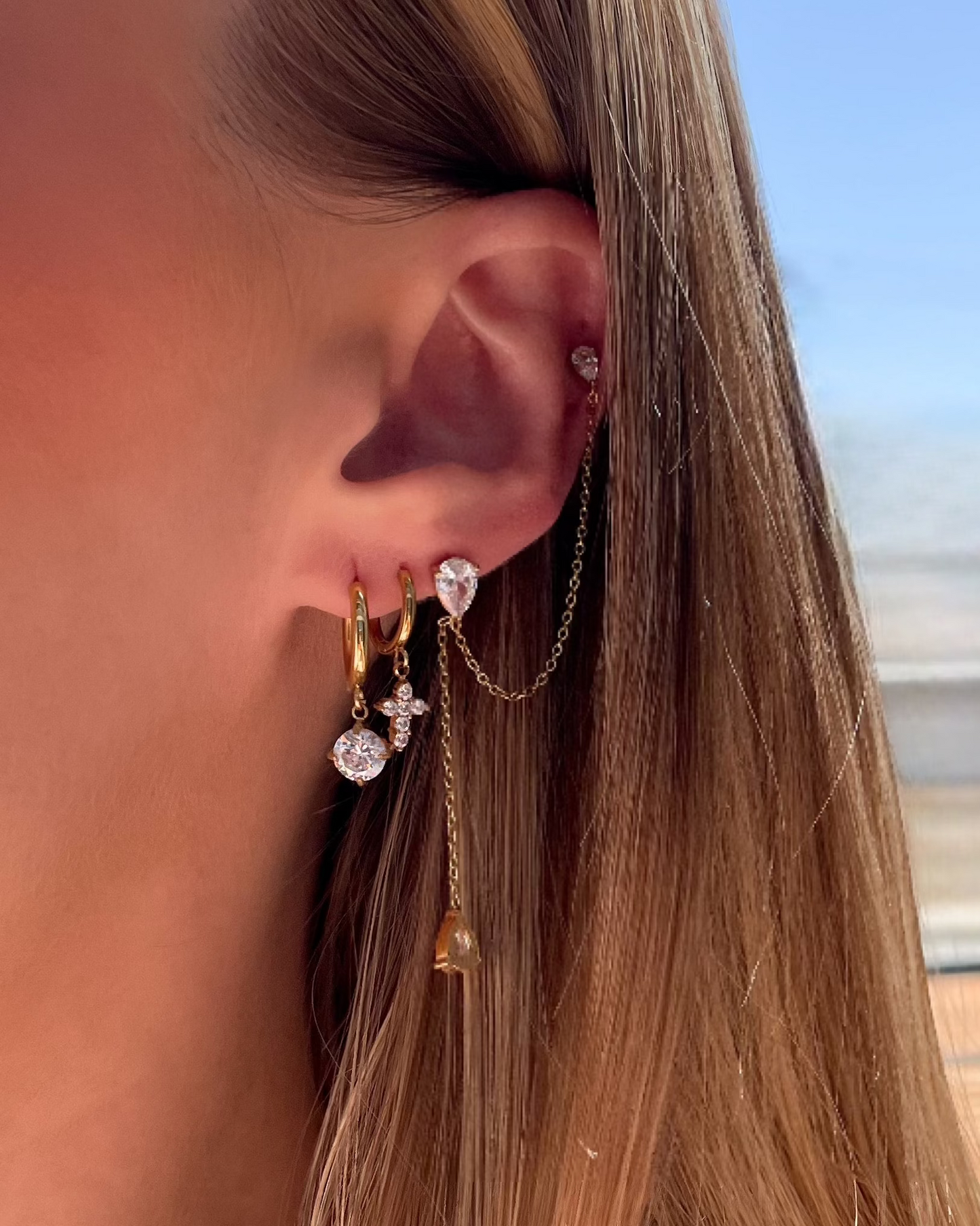 A close-up side profile of a woman's ear wearing multiple gold and silver-toned earrings, including hoop earrings, chain drops, and a small stud, with her blonde hair and a blurred outdoor background visible.