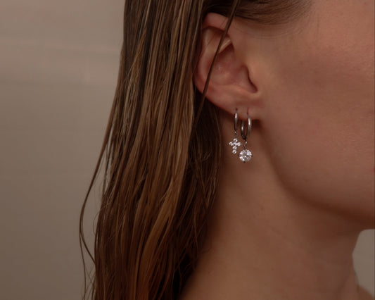 Close-up of a person wearing a silver earring with a clear gemstone against a neutral background