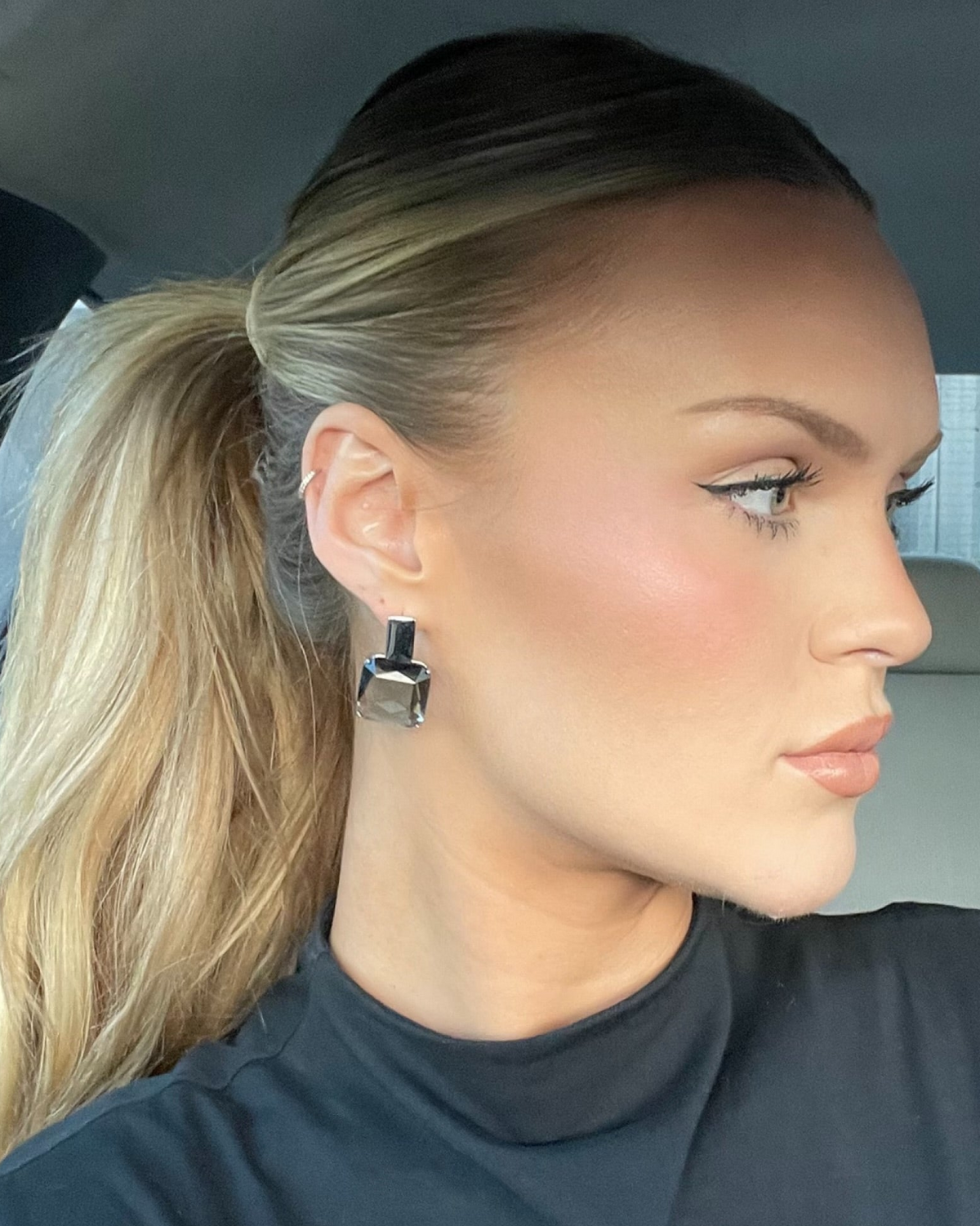 Woman with blonde hair in a ponytail wearing a black turtleneck inside a vehicle.